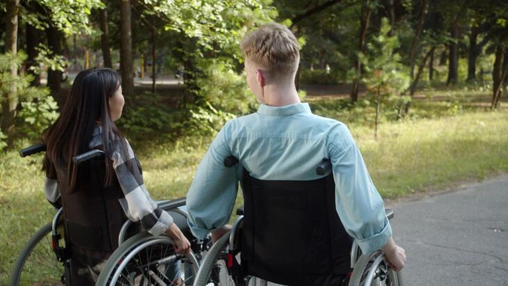 Ung jente og gutt, sitter i hver sin rullestol. Skog/natur.
